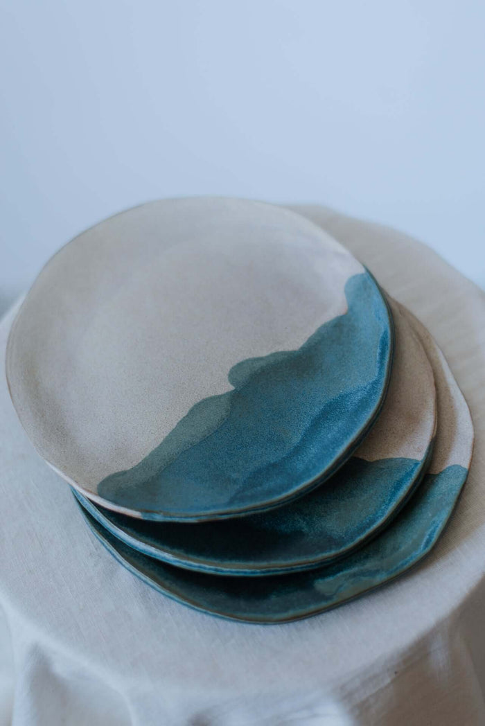 Handcrafted stoneware coupe plate featuring a deep blue wave-like glaze and organic earthy textures.