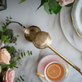 A brass finish tea infuser with a push-open lid, resting on a white cup with tea inside, surrounded by floral decorations.