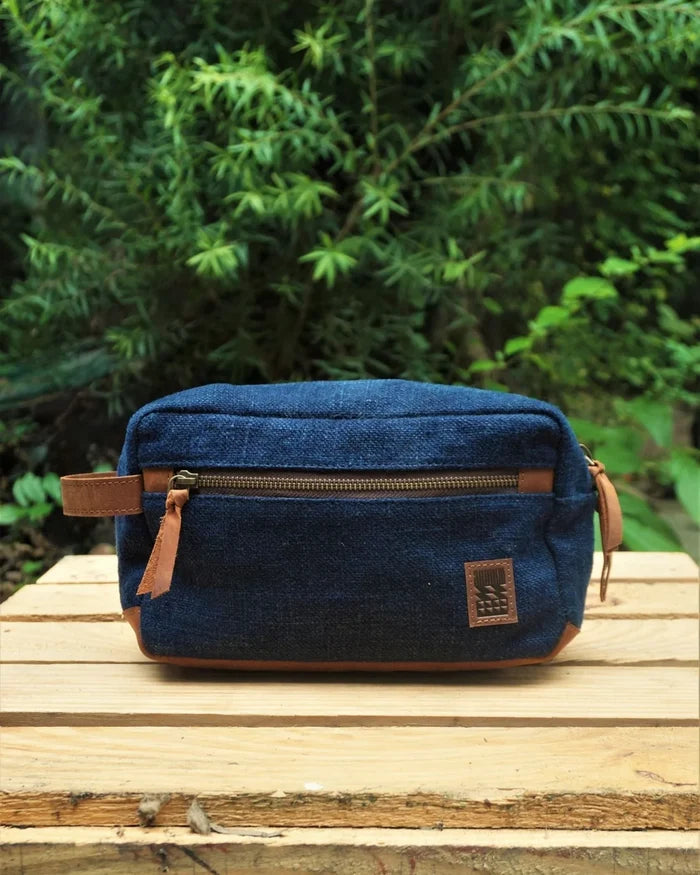 Blue denim toiletry bag with brown leather accents on a wooden surface with green foliage in the background