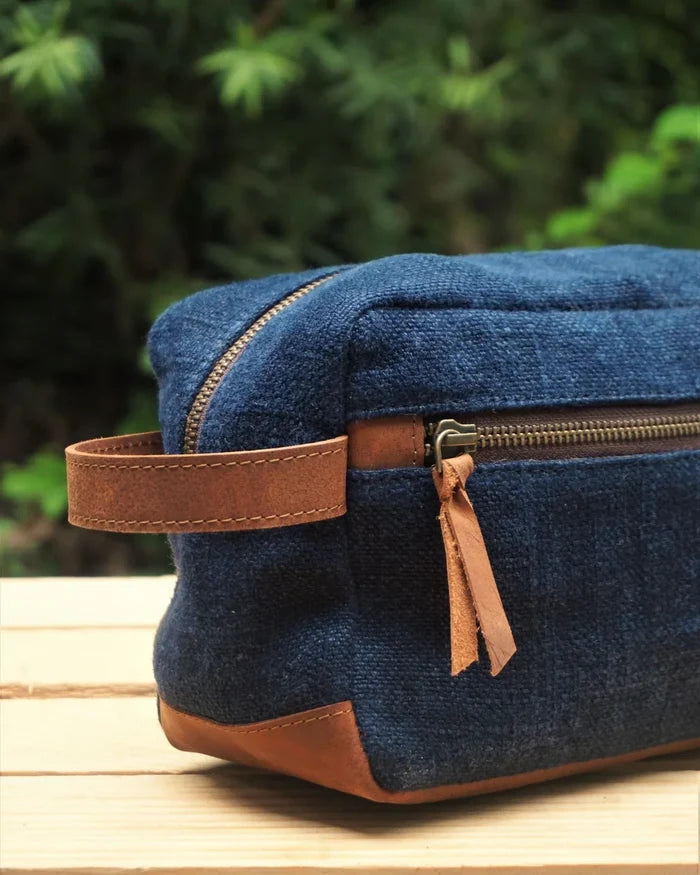 Blue toiletry bag with brown leather strap and zipper on a wooden surface with green foliage in the background