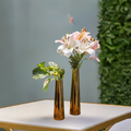 Two gold-finished handcrafted iron vases on a white surface with one tall and one short vase. The tall vase contains a pink and white lily flower arrangement, and the short vase has a green foliage arrangement.