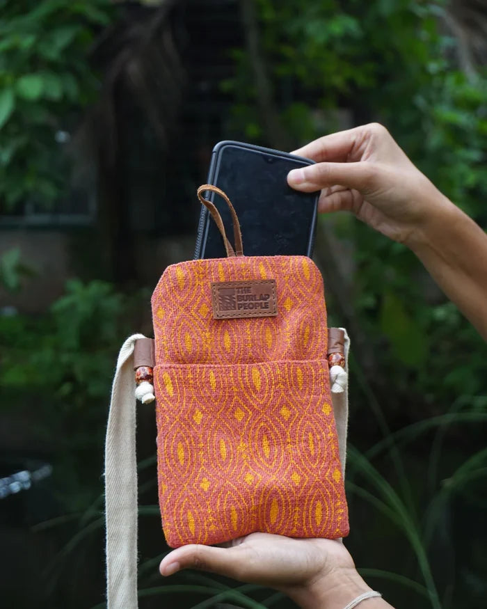 Hand holding an orange patterned phone pouch with a phone inside, against a green leafy background.