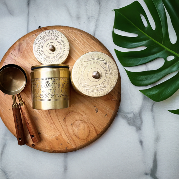 A set of three gold-colored canisters with mandala patterns on them, displayed on a wooden board, accompanied by a wooden spoon and a large green leaf, on a white marble surface.