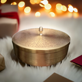 A brass metal spice organizer with a peacock designed engraved and painted lid, sitting on a white surface with bokeh lights in the background.