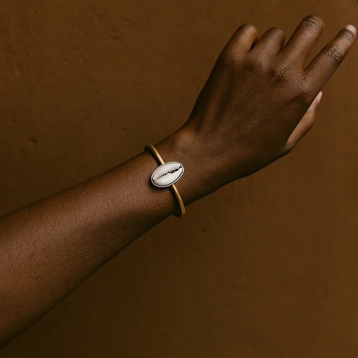 Hand wearing a gold bracelet with a shell charm against a brown background